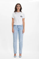 A woman stands facing forward, wearing the GFLApparel 369 Tee in Lite Beam with a small bird design on the chest, light blue jeans, and light blue heels. The background is plain white, subtly hinting at the elegance of sequence 3-6-9 as if channeling the mystical Key to the Universe.