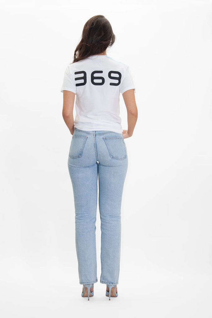 A woman stands facing away from the camera, wearing a white 369 Tee in Lite Beam from GFLApparel with "369" printed on the back and light blue jeans, set against a plain white background. The sequence 3-6-9, often associated with Nikola Tesla's Key to the Universe, subtly adorns her casual attire.