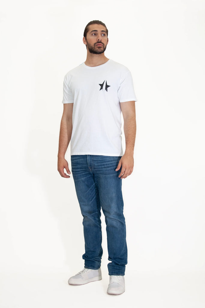 A man with dark hair and a beard stands facing slightly to the left. He wears a white T-shirt with a small black design, blue jeans, and white shoes, against a plain white background, evoking the elegance of GFLApparel's 369 Tee in Lite Beam’s simple yet profound style.