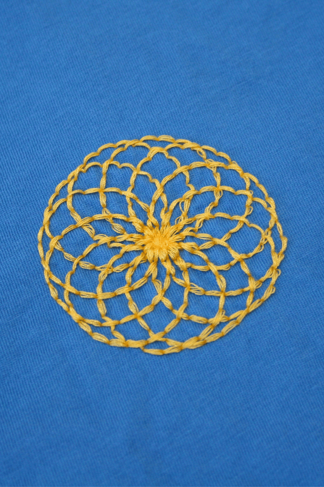 A yellow crocheted doily featuring the Lotus of Life Tee pattern on a blue fabric background by GFLApparel.