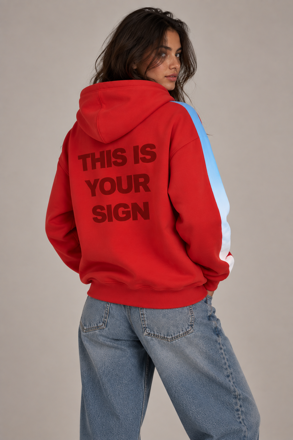 A model wearing a bright red hoodie with 'THIS IS YOUR SIGN' printed in dark redon the back, featuring light blue to white ombre detail on the sleeves, paired with light-wash blue jeans against a neutral beige background.