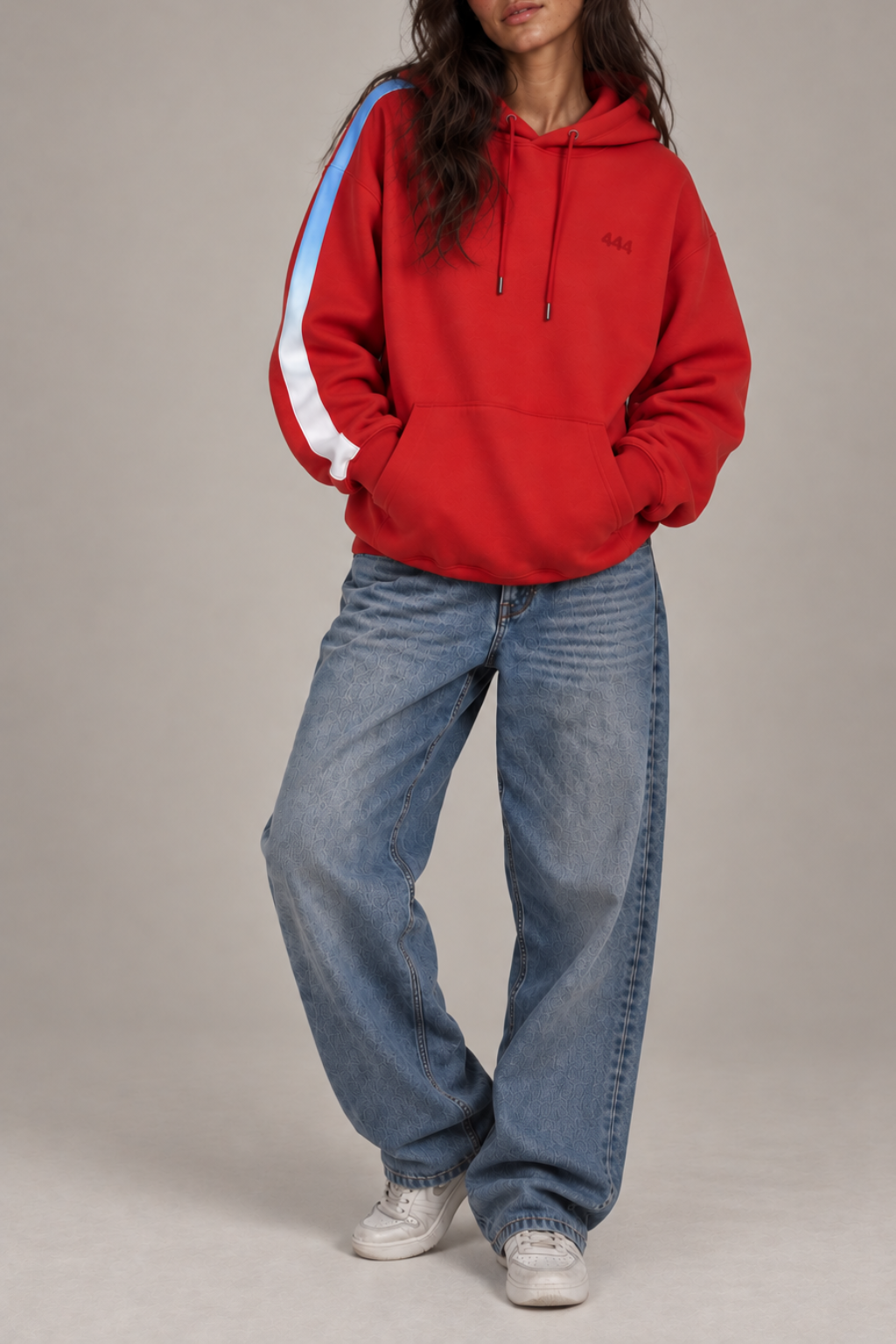 A model wearing a red hoodie with a blue to white ombre stripe on the right sleeve,  paired with wide-leg blue jeans and white sneakers, against a neutral beige studio background.