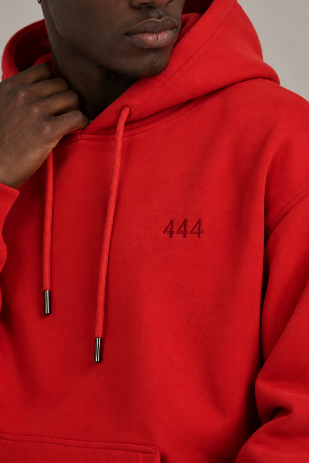 A close-up of a person wearing a bright red pullover hoodie with dark silver drawstrings and dark red '444' embroidered on the left chest, against a neutral light gray background.