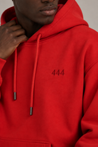 A close-up of a person wearing a bright red pullover hoodie with dark silver drawstrings and dark red '444' embroidered on the left chest, against a neutral light gray background.