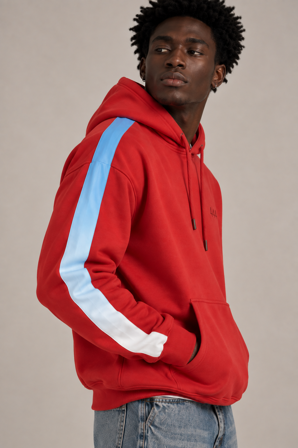 A man wearing a red pullover hoodie with a light blue and white stripe down the sleeve, paired with blue jeans, photographed against a neutral beige studio background.