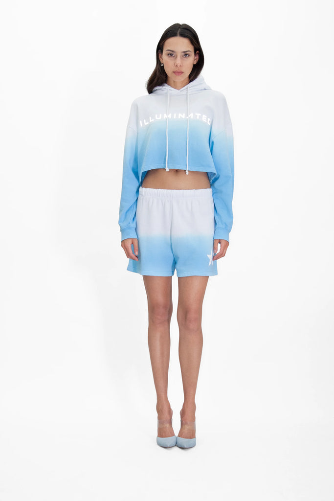 A person stands against a white background wearing a matching blue and white gradient handcrafted hoodie and shorts set paired with light blue heels. The Illuminated Cropped Hoodie in Atmosphere from GFLApparel has the word "ILLUMINATED" printed on it in a reflective print.