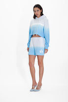 A woman stands against a plain white background wearing an Illuminated Cropped Hoodie in Atmosphere and matching shorts with "ILLUMINATED" in reflective print on the hoodie, paired with light blue high-heeled shoes from GFLApparel.