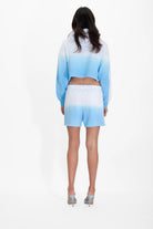 A person stands facing away, wearing the GFLApparel Illuminated Cropped Hoodie in Atmosphere and matching shorts with high heels. The background is plain white.