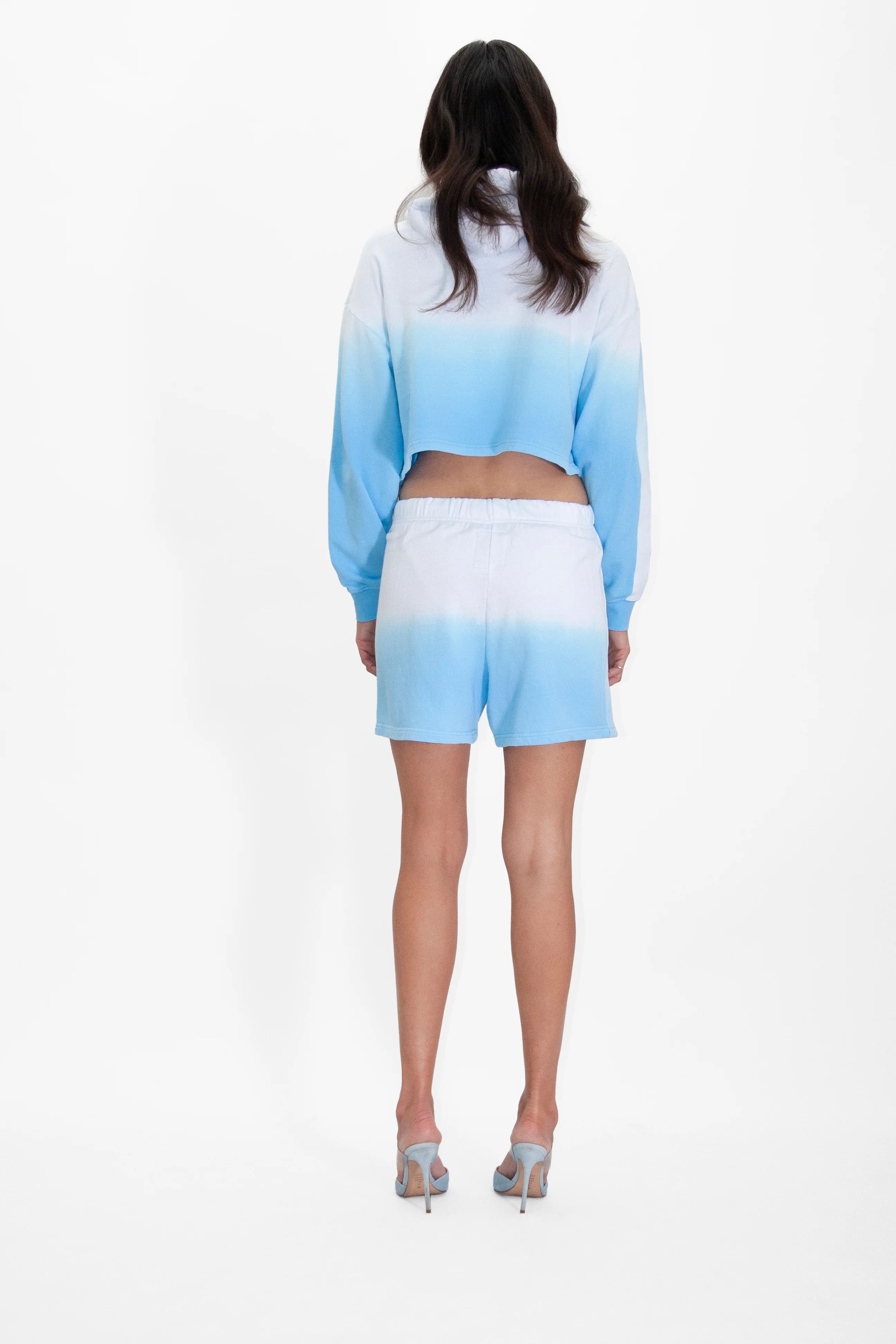 A person stands facing away, wearing the GFLApparel Illuminated Cropped Hoodie in Atmosphere and matching shorts with high heels. The background is plain white.