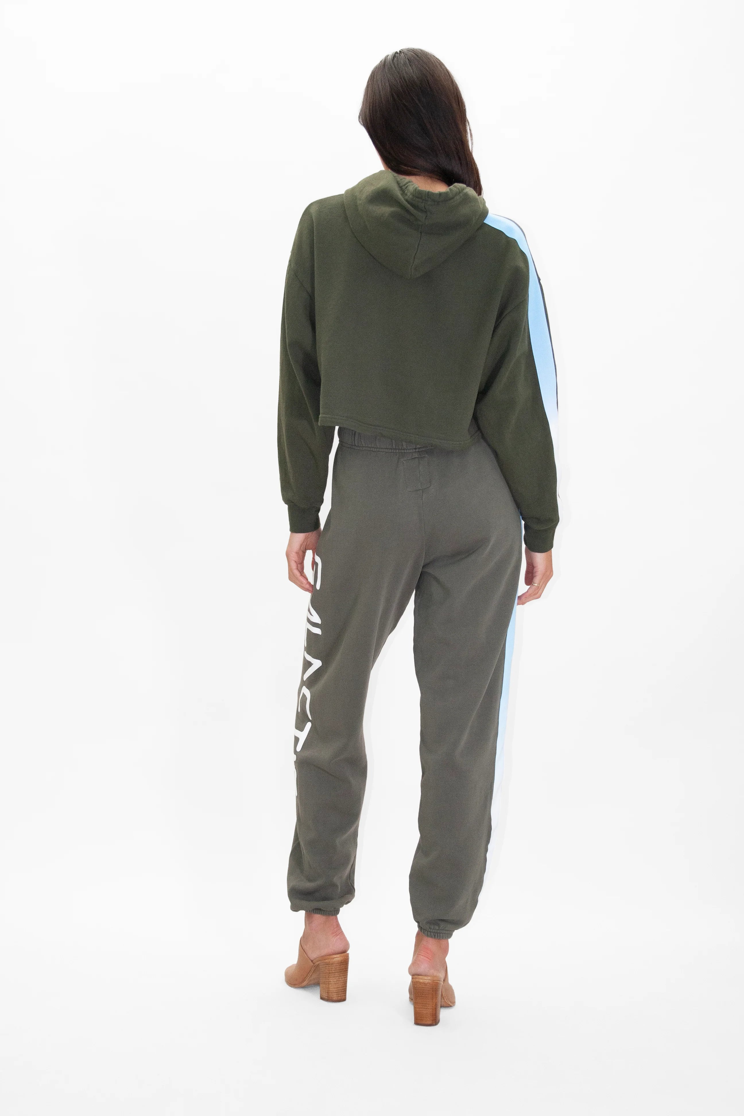 A person stands facing away, wearing a GFLApparel Light Worker Cropped Hoodie in Caladan, matching jogger pants with white lettering on the side, and beige heeled sandals.