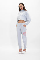 A woman stands against a plain white background, wearing a light grey, two-piece outfit consisting of the GFLApparel Merkaba Cropped Hoodie in Galactic Gray and joggers with sacred geometry designs, and white high-heeled shoes.