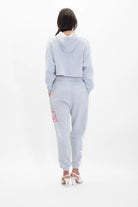 A person with long dark hair wearing a light gray **Merkaba Cropped Hoodie in Galactic Gray** by **GFLApparel** and matching jogger pants with red and white Sacred Geometry designs on the left leg, stands facing away. They are also wearing white high-heeled shoes.