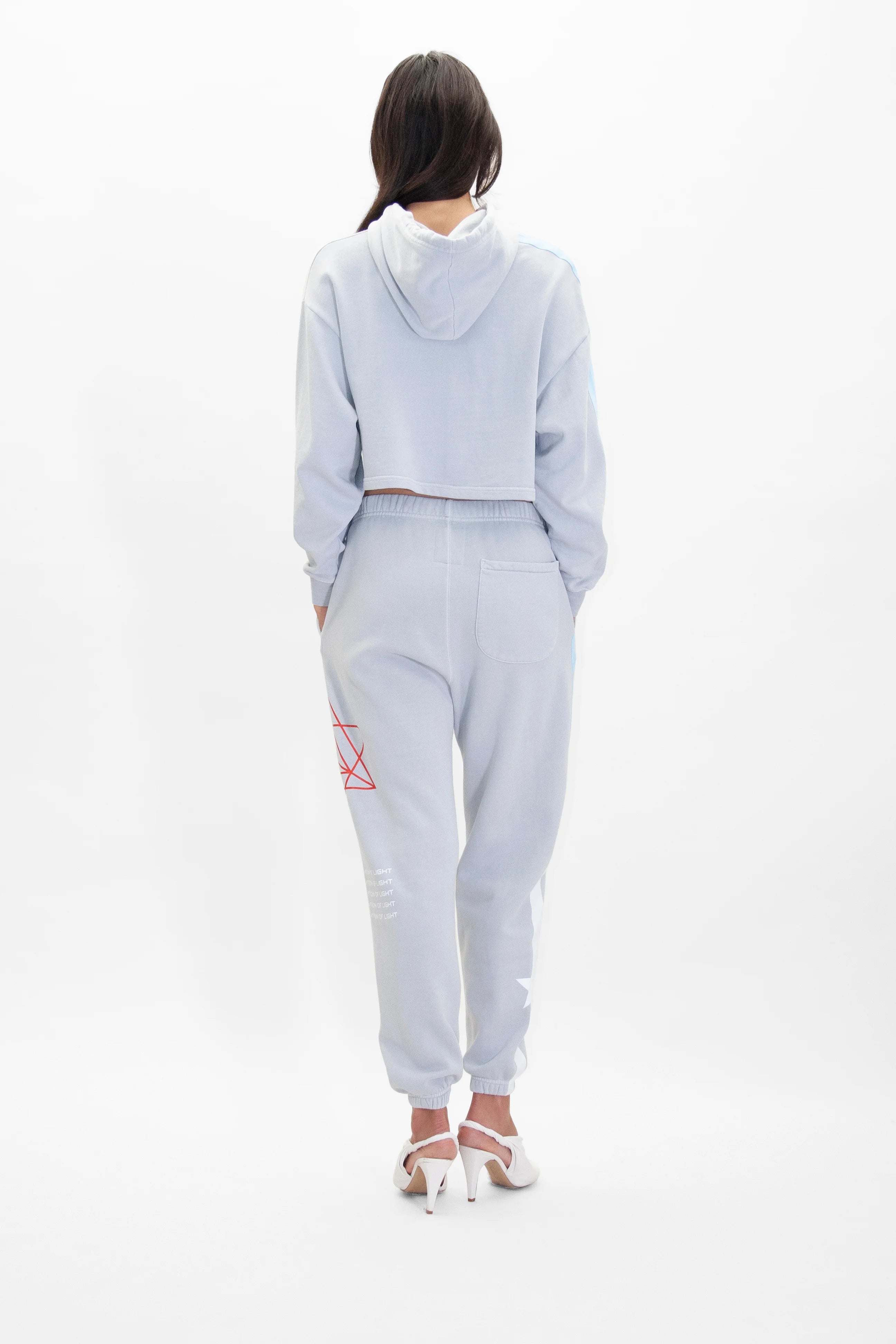 A person with long dark hair wearing a light gray **Merkaba Cropped Hoodie in Galactic Gray** by **GFLApparel** and matching jogger pants with red and white Sacred Geometry designs on the left leg, stands facing away. They are also wearing white high-heeled shoes.