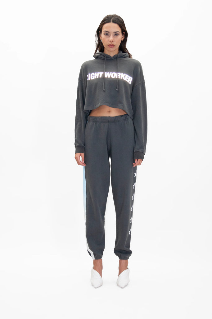 A person stands against a plain white background wearing a dark gray Light Worker Cropped Hoodie in Space Glow by GFLApparel with "LIGHT WORKER" printed in reflective print, matching sweatpants with white side stripes, and white shoes.