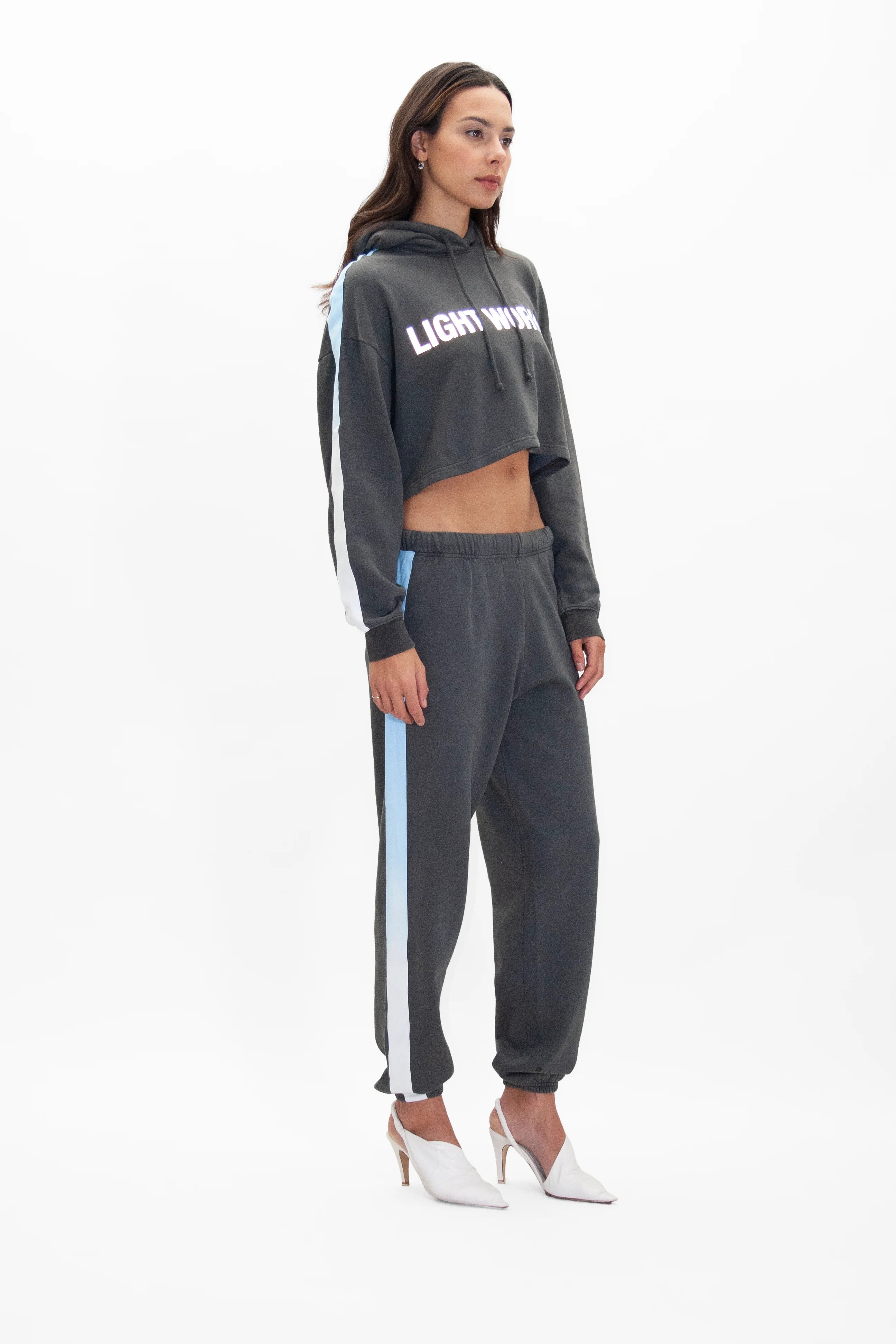 A woman stands in profile, wearing the GFLApparel Light Worker Cropped Hoodie in Space Glow with the text "LIGHT WAVE" in reflective print and matching joggers with light blue side stripes. She is also wearing white high heels.