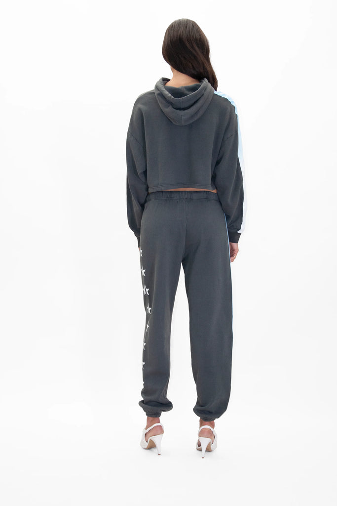 A person is standing with their back to the camera, wearing a Light Worker Cropped Hoodie in Space Glow from GFLApparel and joggers with reflective star designs on the side, paired with white high-heeled shoes.