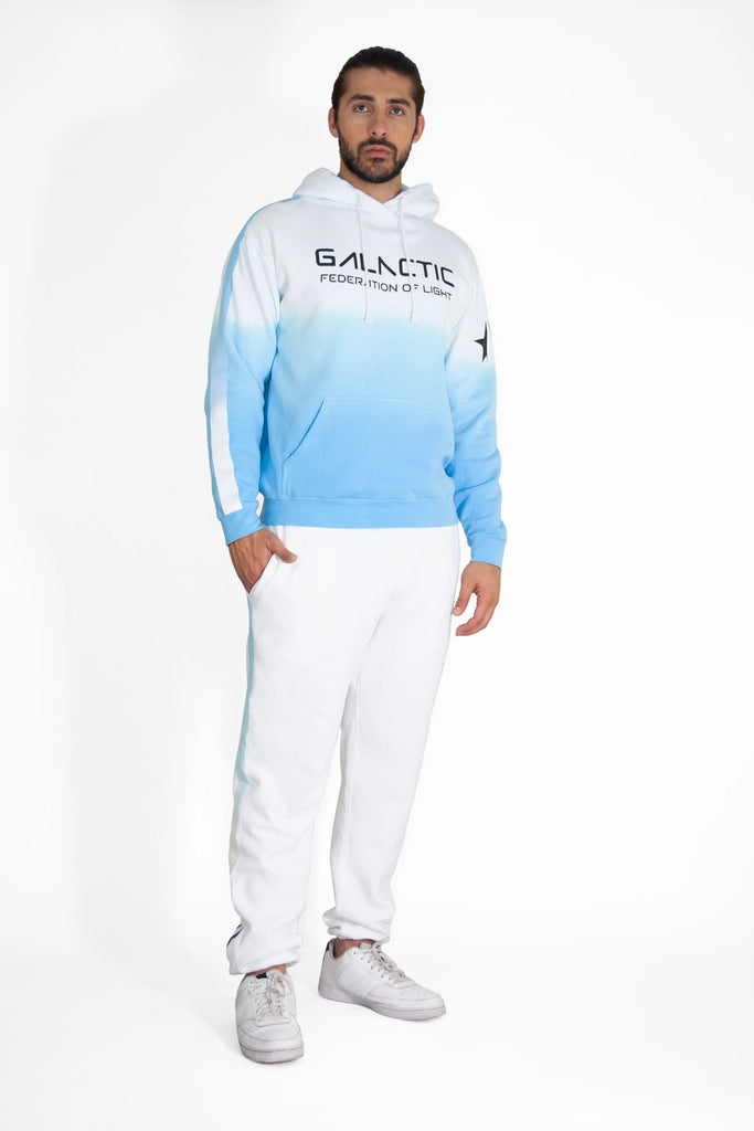 A man stands against a white background wearing a Galactic Hoodie in Atmosphere by GFLApparel, featuring the "Galactic Federation of Light" emblem. Paired with cozy cotton blend white pants and matching sneakers, he exudes comfort and style.