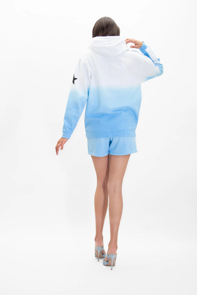 A person is standing with their back to the camera, wearing a GFLApparel Galactic Hoodie in Atmosphere with an ombré dye of white and blue, paired with blue shorts and light blue high heels.
