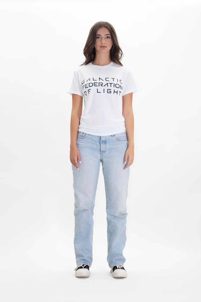 A woman stands against a plain white background, wearing a white GFLApparel GFL Stack Tee in Lite Beam and light blue jeans. She has long dark hair and is facing forward.