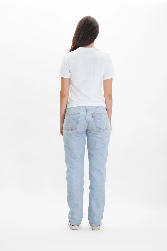 A person with long hair wearing a white GFLApparel GFL Stack Tee in Lite Beam, blue jeans, and beige shoes is standing with their back facing the camera against a white background.