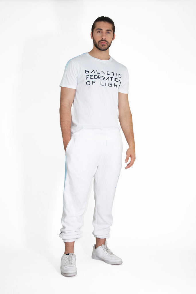 A man wearing a white GFLApparel GFL Stack Tee in Lite Beam featuring the text "Galactic Federation of Light" and matching white sweatpants stands against a plain white background.