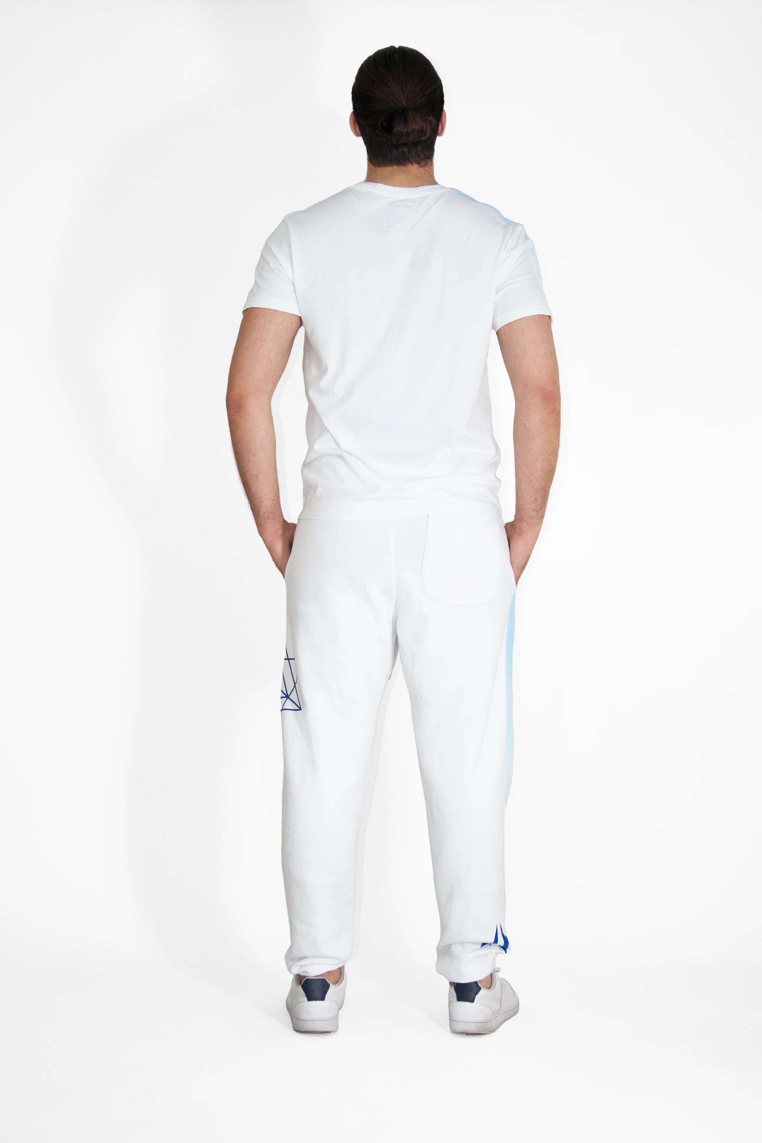 A person with long hair tied back is standing with their back to the camera, wearing a white GFL Stack Tee in Lite Beam by GFLApparel, white pants with blue designs, and white shoes.