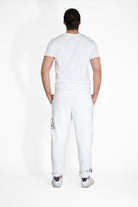 A person with long hair tied back is standing with their back to the camera, wearing a white GFL Stack Tee in Lite Beam by GFLApparel, white pants with blue designs, and white shoes.