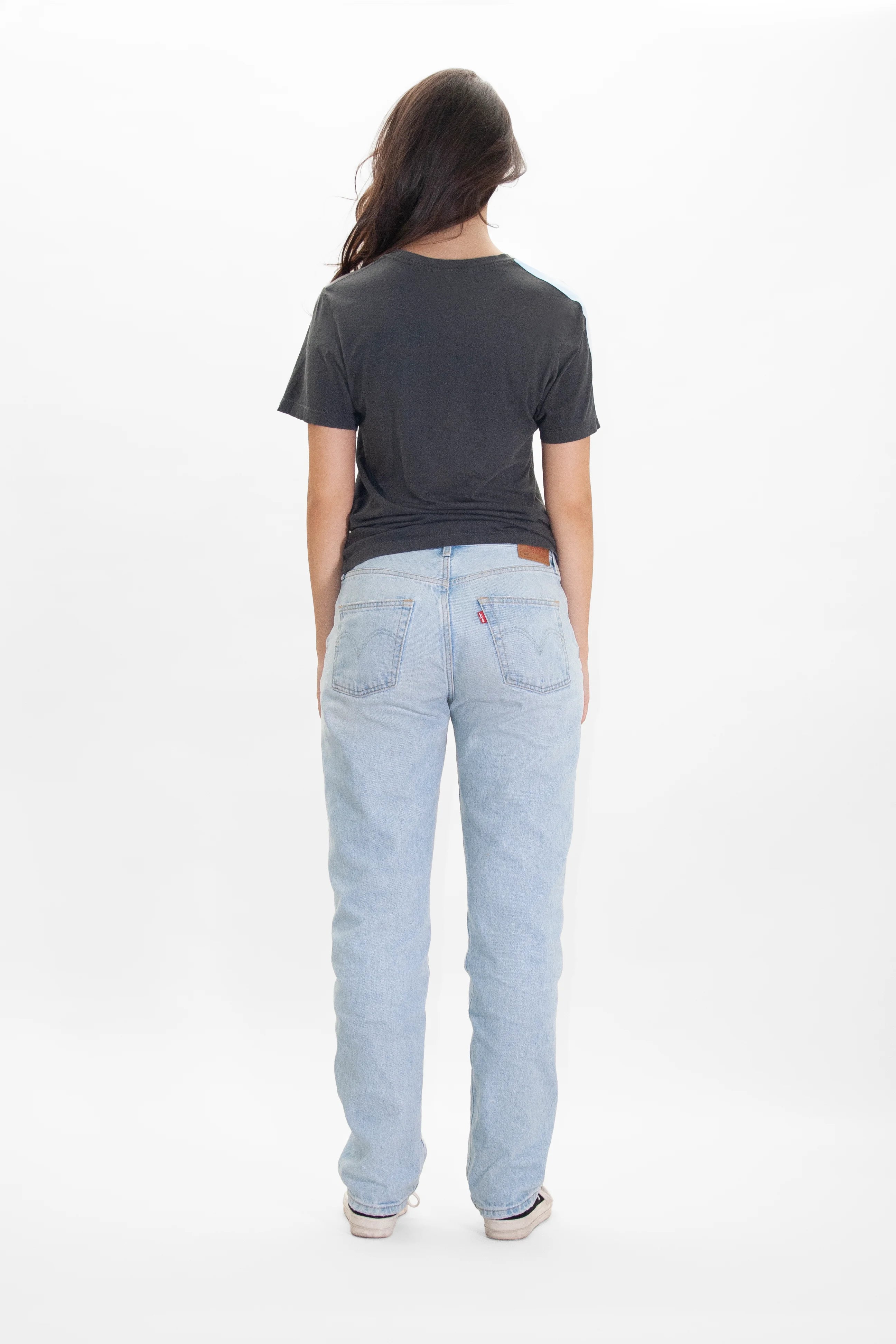 A person wearing a dark grey GFL Stack Tee in Space Glow from GFLApparel and light blue jeans, standing and facing away from the camera against a plain white background.