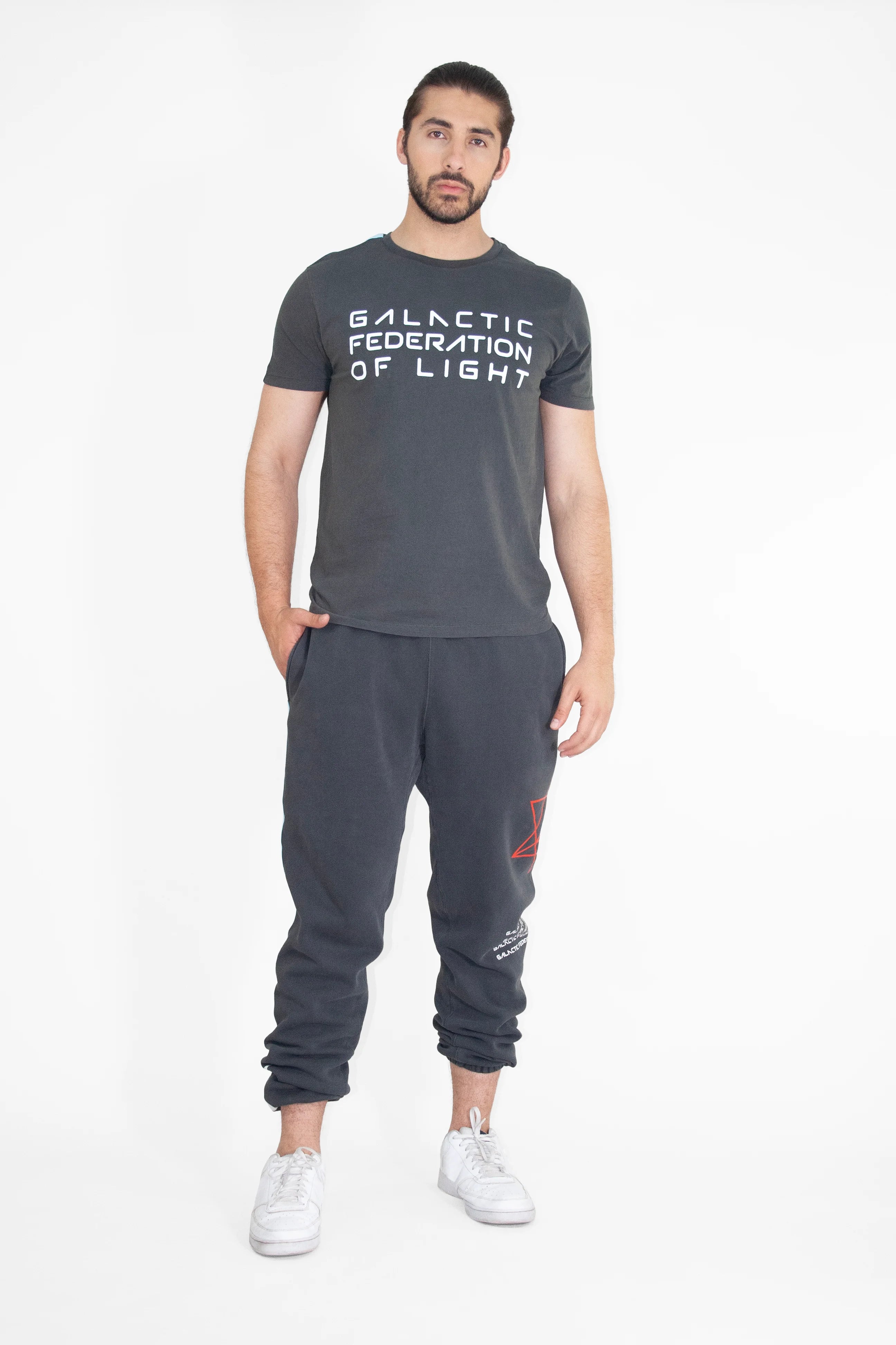 A man stands in a relaxed posture wearing a GFLApparel GFL Stack Tee in Space Glow, dark jogger pants, and white sneakers against a plain white background.