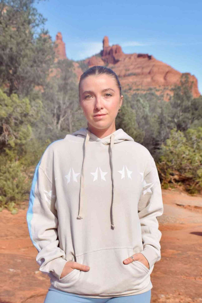 A woman in a GFLApparel STARS HOODIE IN DUNE stands in front of a scenic red rock formation under a clear blue sky.