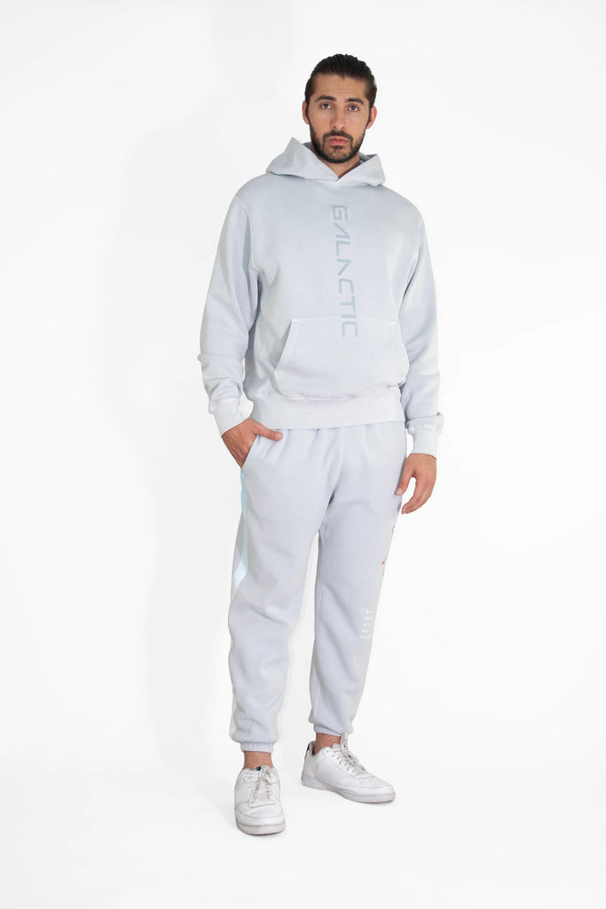 A man wearing a GFLApparel Hypergalactic Hoodie in Galactic Gray made of heavyweight cotton blend, paired with matching jogger pants, stands against a white background. He is also wearing white sneakers.