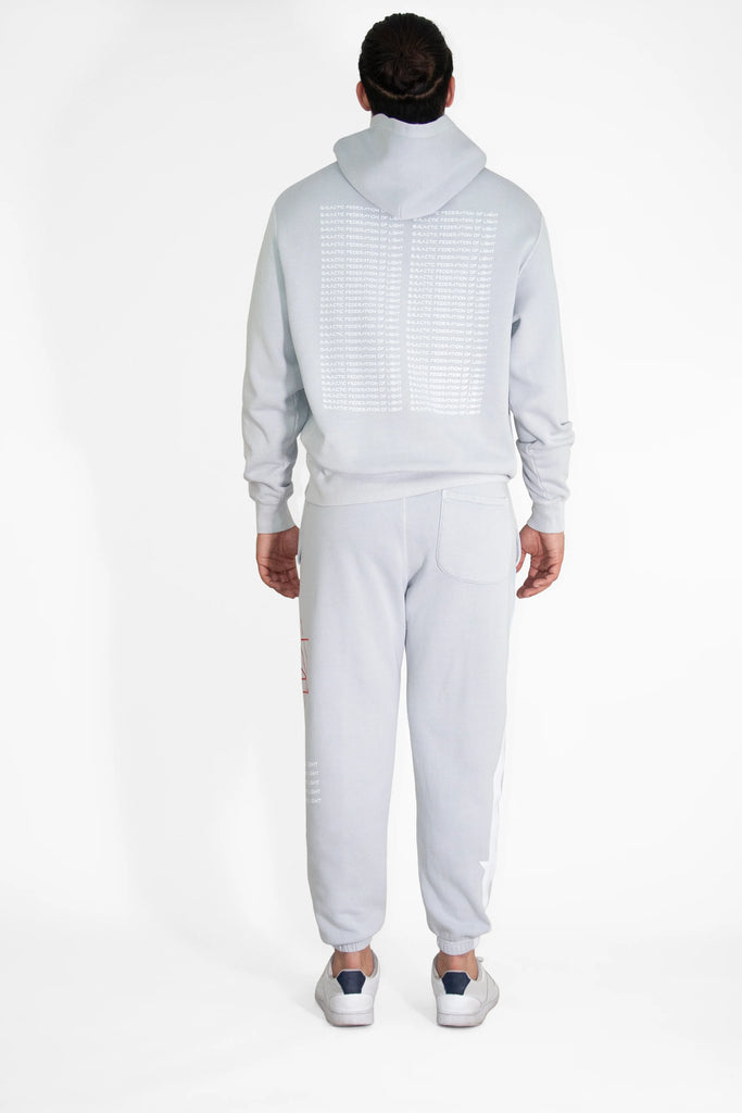 A person is standing with their back facing the camera, wearing a light gray Hypergalactic Hoodie in Galactic Gray by GFLApparel made from a heavyweight cotton blend and matching sweatpants with white sneakers. The hoodie, handcrafted in LA, has text printed on the back.