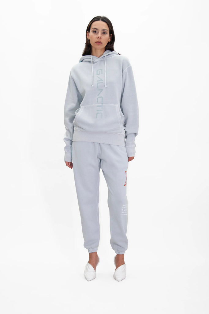 A person stands against a white background wearing a light gray Hypergalactic Hoodie in Galactic Gray from GFLApparel, crafted from heavyweight cotton blend with "GALACTIC" on it and matching jogger pants, along with white slip-on shoes.