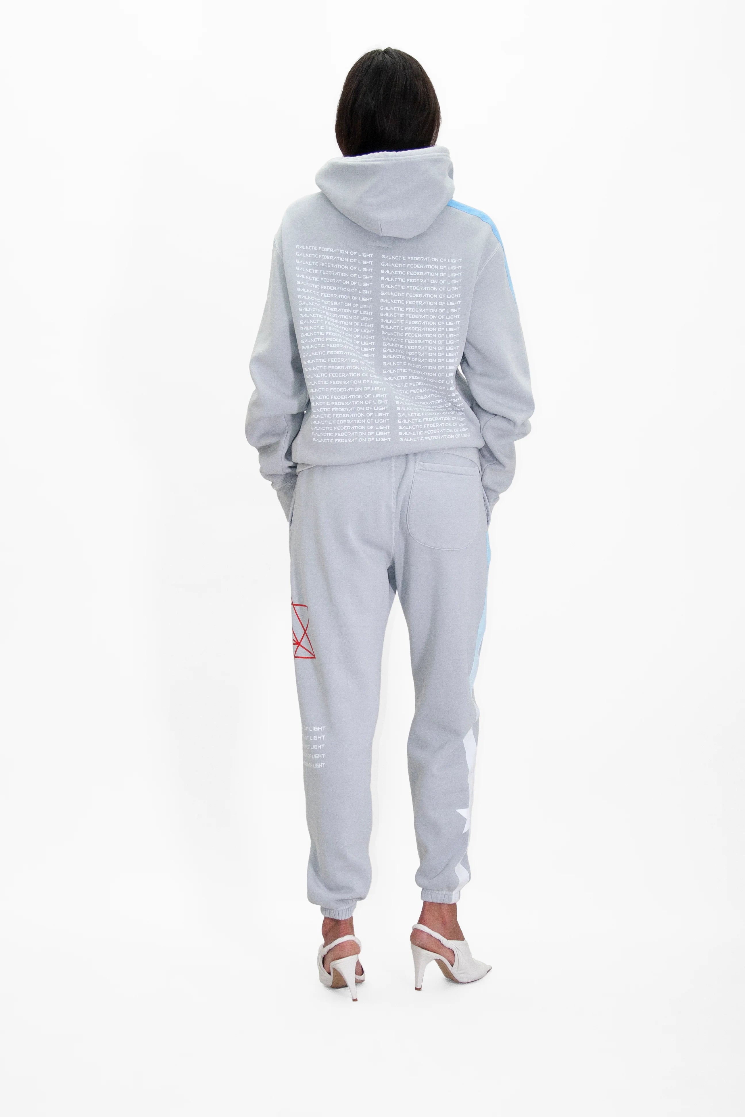 A person stands facing away, wearing a light gray hoodie and joggers with various white and red graphic designs. The Hypergalactic Hoodie in Galactic Gray by GFLApparel, handcrafted in LA from a heavyweight cotton blend, is paired with chic white high-heeled shoes.