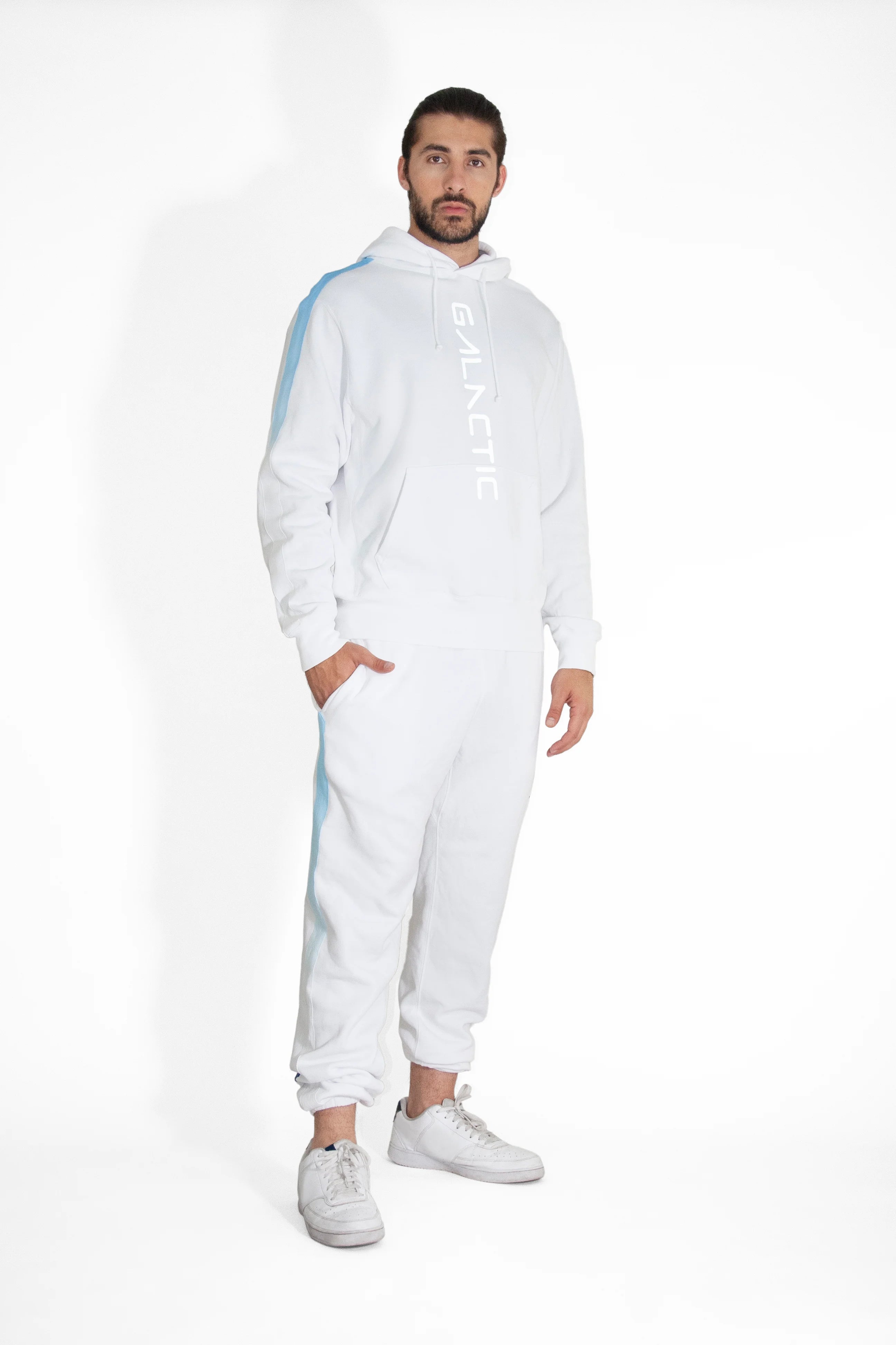 A person stands wearing a white Hypergalactic Hoodie in Lite Beam by GFLApparel, handcrafted in LA, with the word "GALACTIC" printed on it. They match it with white sweatpants and sneakers against a plain white background.