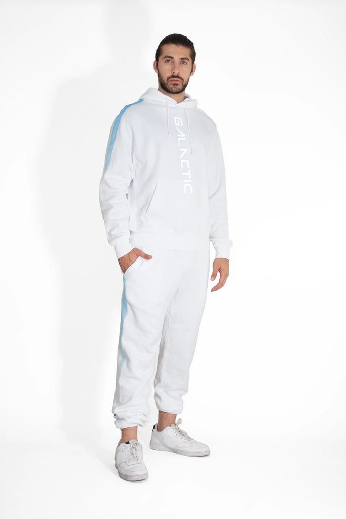 A person stands wearing a white Hypergalactic Hoodie in Lite Beam by GFLApparel, handcrafted in LA, with the word "GALACTIC" printed on it. They match it with white sweatpants and sneakers against a plain white background.
