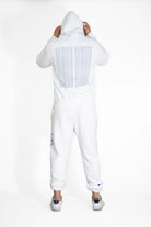 Person wearing a GFLApparel Hypergalactic Hoodie in Lite Beam, made from a heavyweight cotton blend, and white pants with text on the back, standing with their back to the camera, holding the hood. This unique piece is handcrafted in LA.