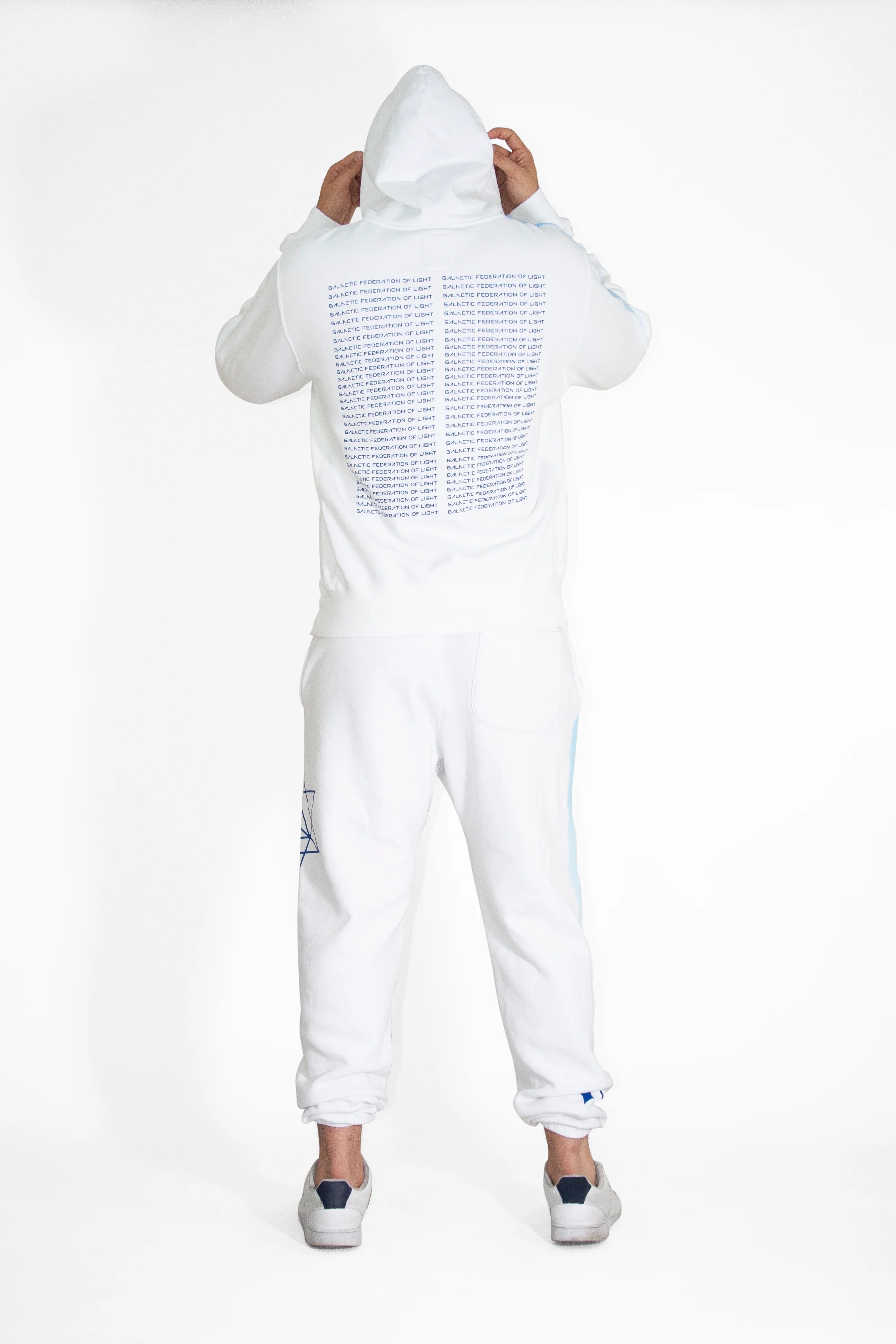 Person wearing a GFLApparel Hypergalactic Hoodie in Lite Beam, made from a heavyweight cotton blend, and white pants with text on the back, standing with their back to the camera, holding the hood. This unique piece is handcrafted in LA.