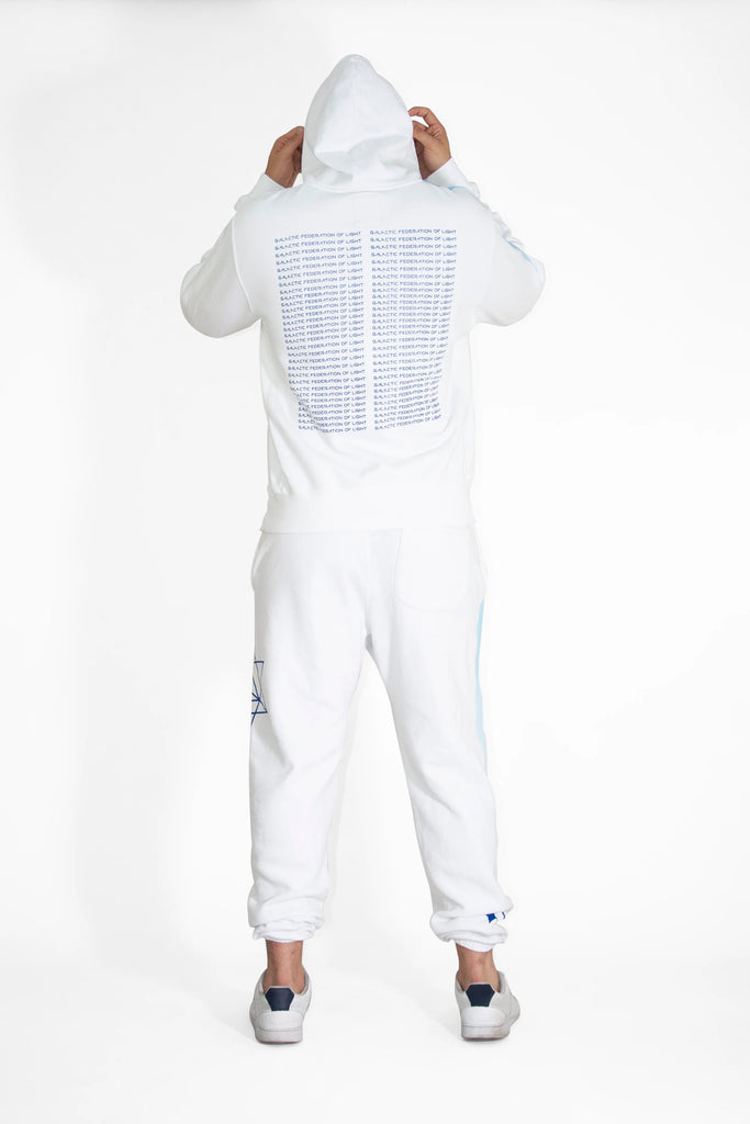 Person wearing a GFLApparel Hypergalactic Hoodie in Lite Beam, made from a heavyweight cotton blend, and white pants with text on the back, standing with their back to the camera, holding the hood. This unique piece is handcrafted in LA.
