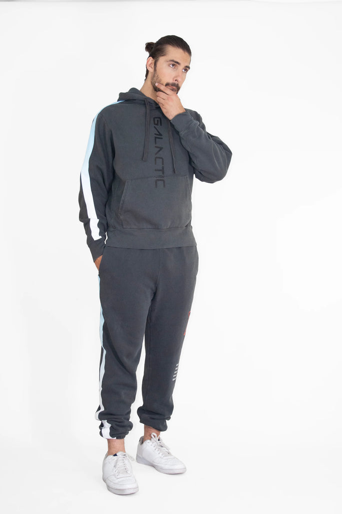 A man with tied-back hair is dressed in a dark sweatsuit made from a heavyweight cotton blend with white and light blue stripes, featuring the word "Hypergalactic Hoodie in Space Glow" on the front. He is standing thoughtfully with his hand on his chin. The product is from GFLApparel.