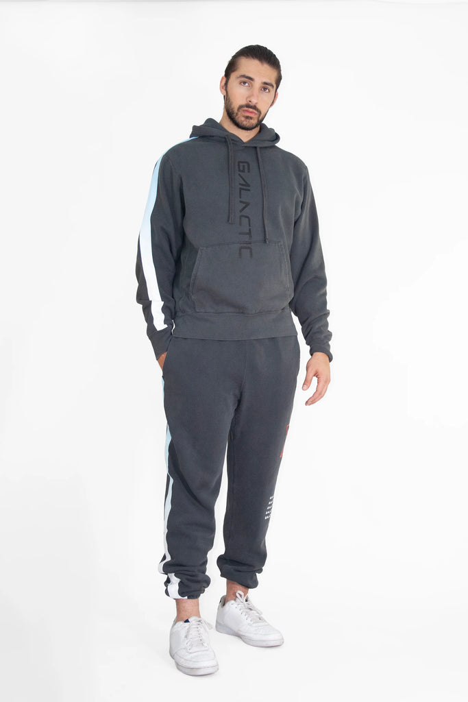A man wearing a dark hoodie labeled "Hypergalactic Hoodie in Space Glow" by GFLApparel on the front pocket stands against a plain, white background. The handcrafted in LA outfit includes matching dark sweatpants and white sneakers, all made from a heavyweight cotton blend for ultimate comfort.