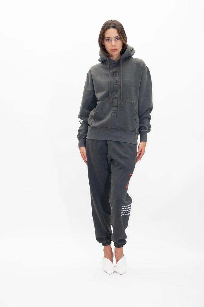 A person is wearing a dark grey Hypergalactic Hoodie in Space Glow from GFLApparel, handcrafted in LA from heavyweight cotton blend fabric, with matching sweatpants, standing against a plain white background.
