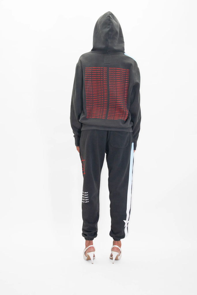 A person wearing a GFLApparel Hypergalactic Hoodie in Space Glow made from heavyweight cotton blend, featuring red text on the back, stands facing away in matching black pants with white and blue side stripes and white heeled shoes.