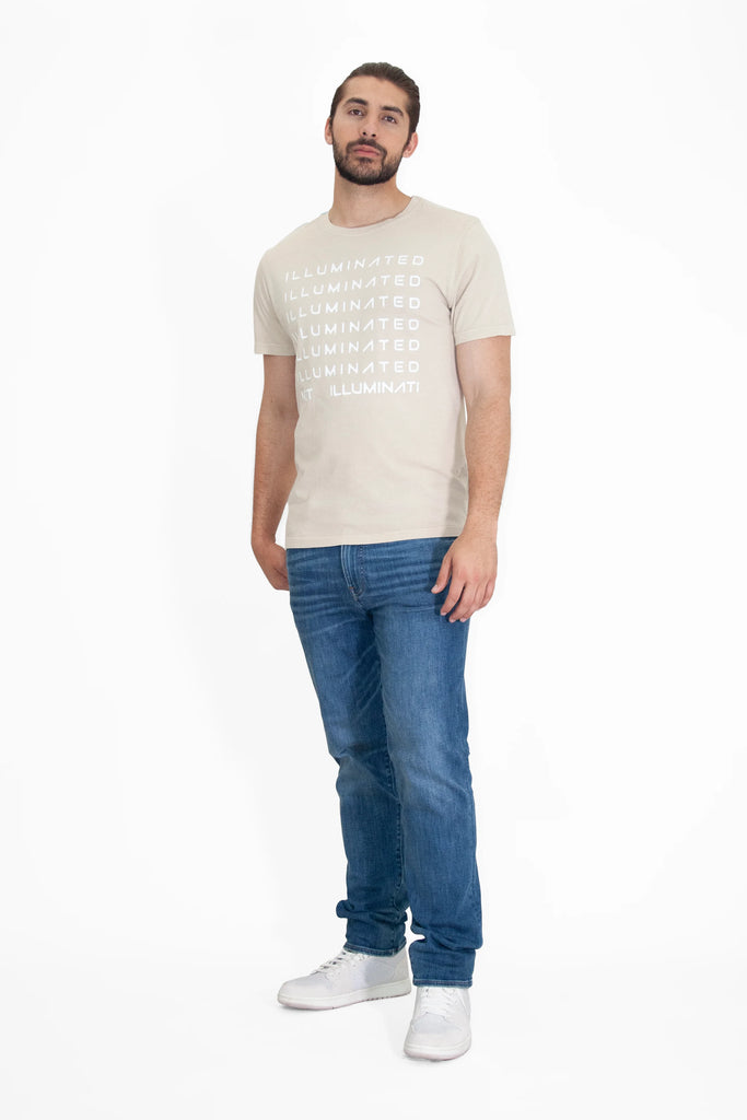 A man stands against a white background wearing a light-colored, soft, and breathable cotton GFLApparel Illuminated Tee in Dune with the word "ILLUMINATED" repeated in bold print, paired with blue jeans and white sneakers.