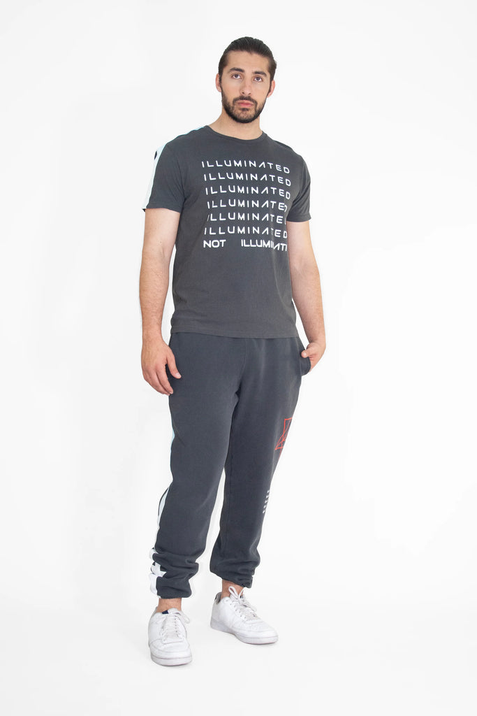 A man with a beard wearing a dark gray, breathable cotton Illuminated Tee in Space Glow from GFLApparel with the word "ILLUMINATED" repeated in bold print and "NOT ILLUMINATED" at the bottom, paired with dark sweatpants and white sneakers, standing against a white background.
