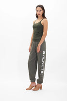 A woman stands wearing a green tank top and gray, super soft Galactic Sweatpants in Caladan by GFLApparel with the word "GALACTIC" printed on the leg, paired with brown heeled sandals against a plain white background.