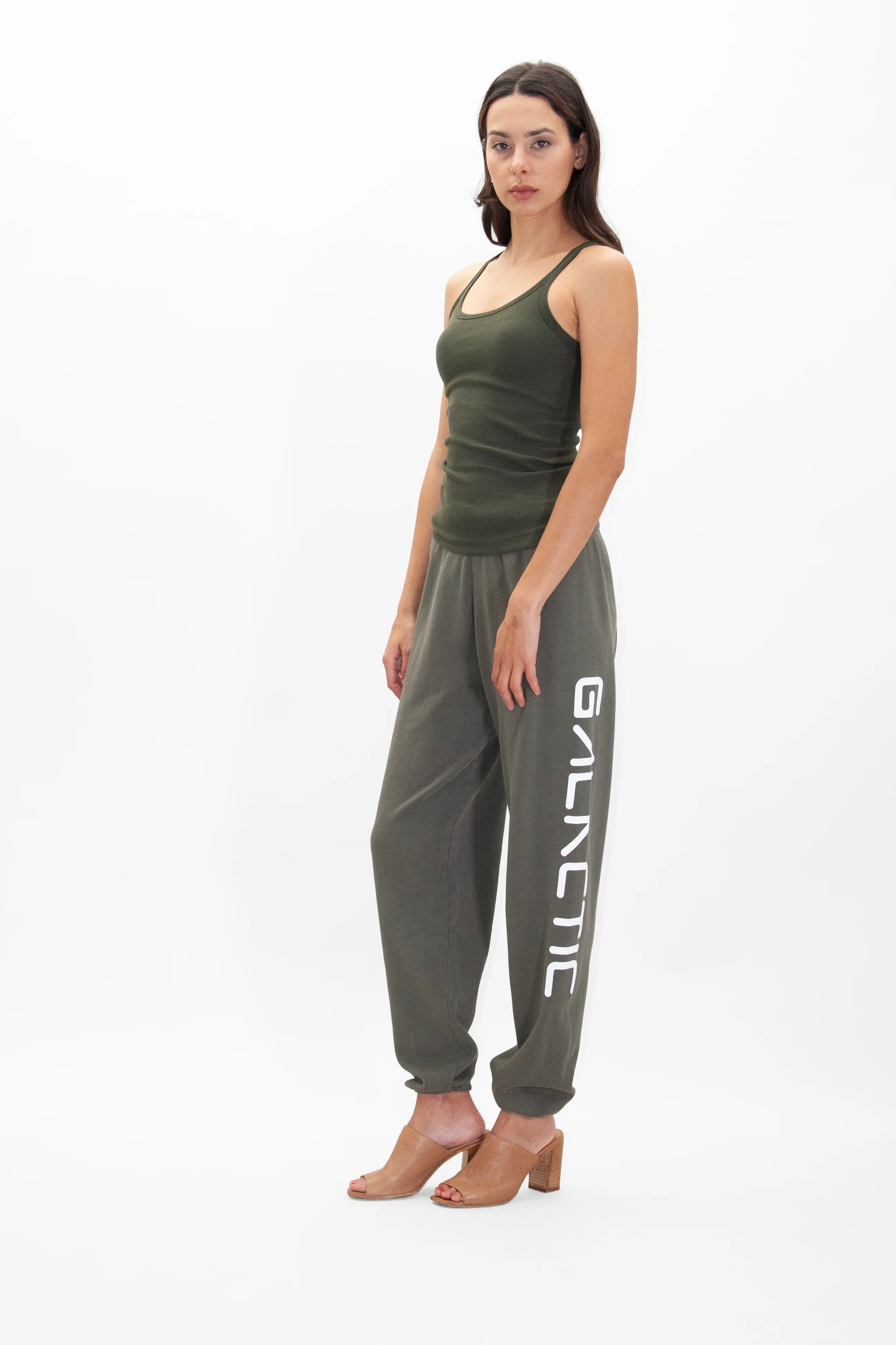 A woman stands wearing a green tank top and gray, super soft Galactic Sweatpants in Caladan by GFLApparel with the word "GALACTIC" printed on the leg, paired with brown heeled sandals against a plain white background.