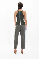 A woman is standing facing away, wearing a dark green tank top with a vertical light blue stripe, Galactic Sweatpants in Caladan by GFLApparel in dark green with white text on the side and a light blue stripe, and tan high-heeled shoes.