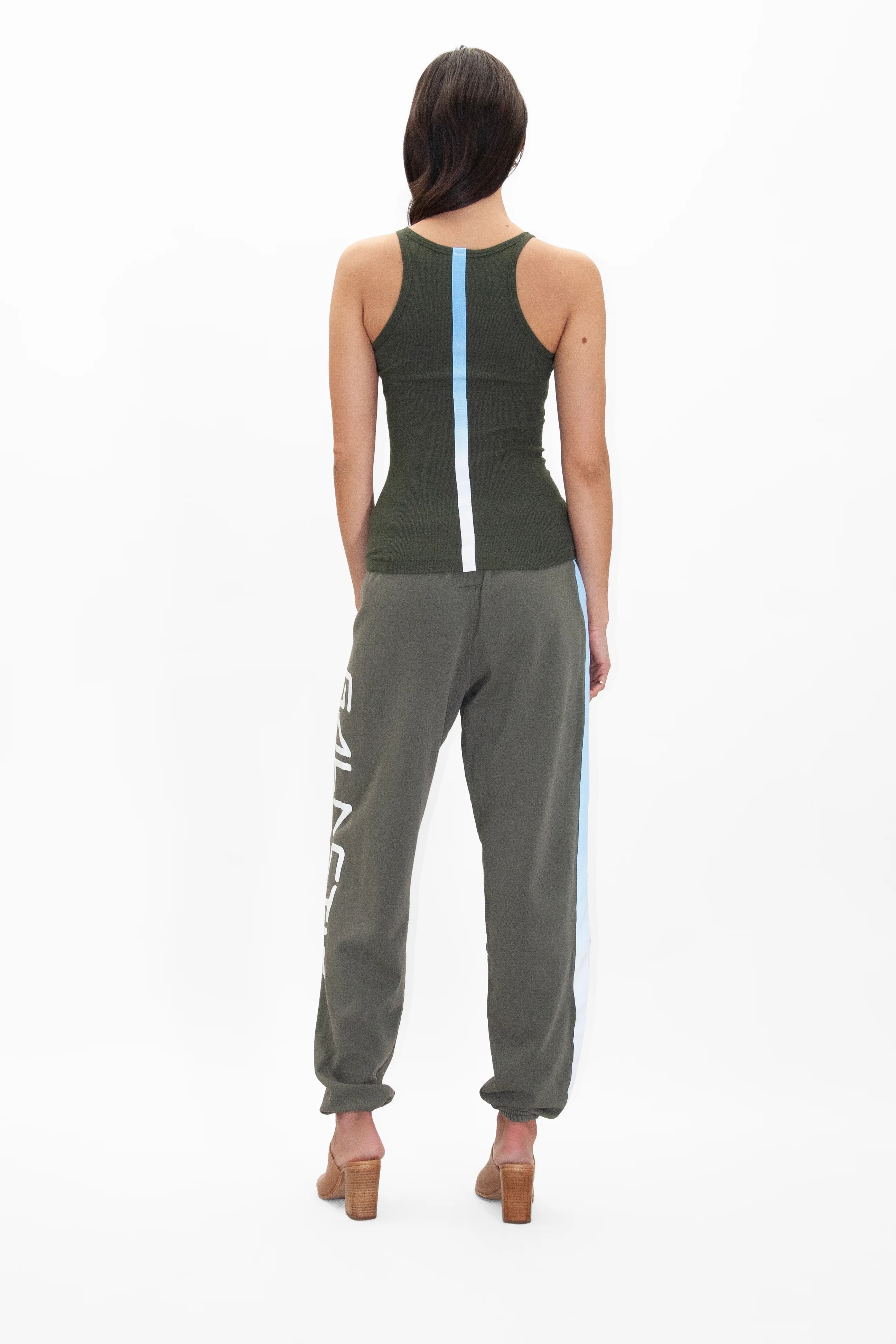 A woman is standing facing away, wearing a dark green tank top with a vertical light blue stripe, Galactic Sweatpants in Caladan by GFLApparel in dark green with white text on the side and a light blue stripe, and tan high-heeled shoes.