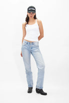 A person stands against a plain white background wearing a white tank top, light blue jeans, black boots, and a must-have Light Worker Baseball Cap by GFLApparel.